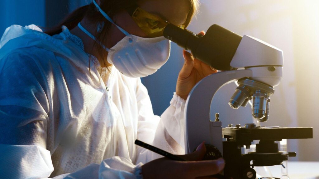 Female chemist in protective workwear looking through the microscope and drug testing